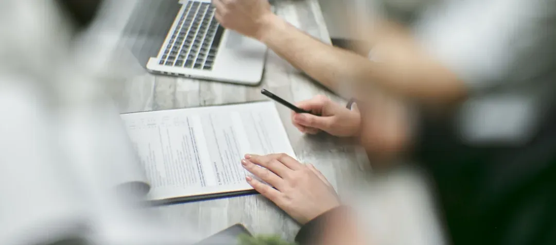 Manos revisando documentos junto a un portátil en una mesa de reuniones